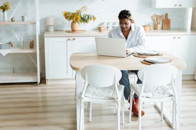 A woman typing on her laptop at a dining table in a bright and modern kitchen setting.