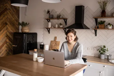 Cheerful female in casual clothes sitting at table with laptop and bottle of milk while browsing internet on laptop during free time at home and smiling looking at camera