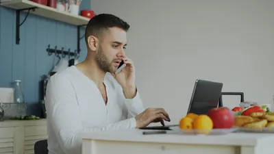 Man multitasking with laptop and phone at home, surrounded by fruit and a modern kitchen.