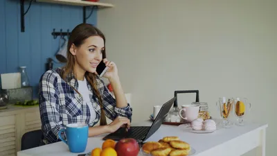 Young woman multitasking on phone and laptop in a cozy kitchen setting.