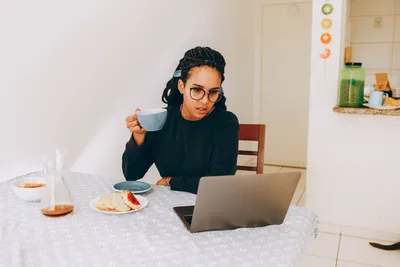 Young woman enjoying coffee while working remotely on her laptop at home kitchen table.